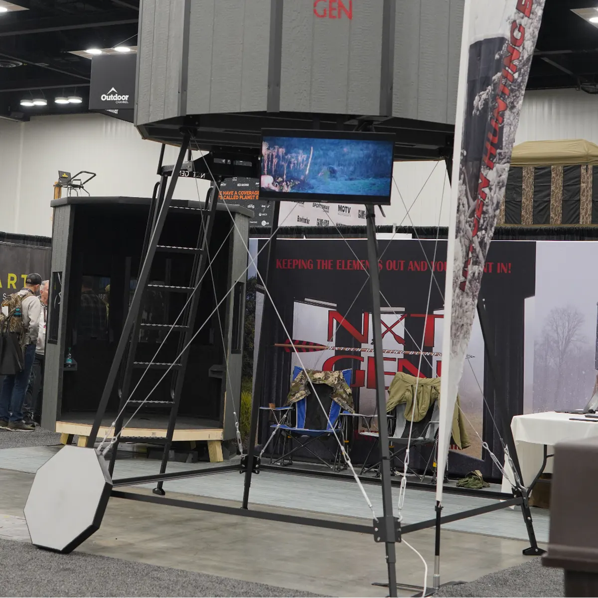 A NXTGen blind on a tall aluminum tower at a trade show, with a second blind on the floor beside it.